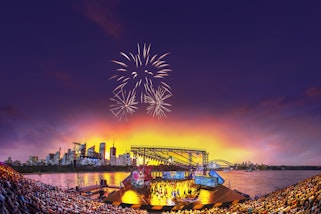 A large seated audience watch performers on a stage on water with fireworks and  skycrapers behind