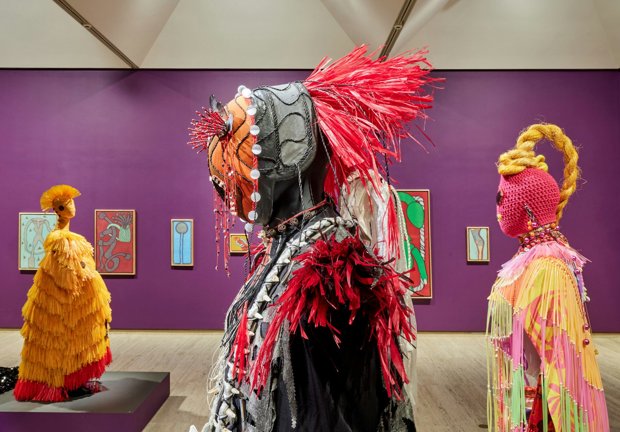 Sculptural figures in colourful costumes stand in a gallery space with paintings hanging on a purple wall