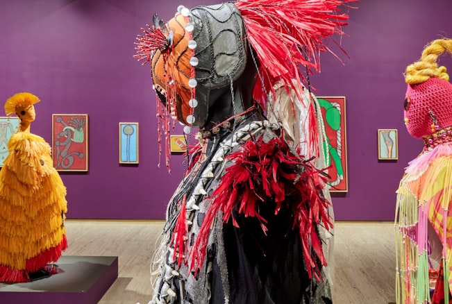 Sculptural figures in colourful costumes stand in a gallery space with paintings hanging on a purple wall