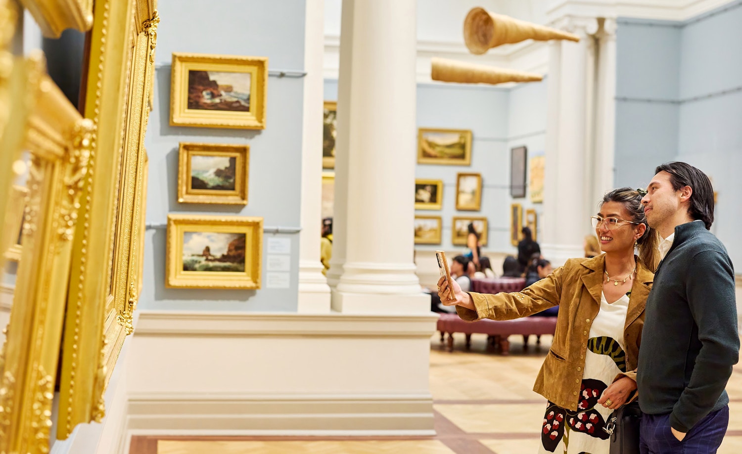 Two people look at gold-framed artworks in a gallery space hung with paintings and two horn-like suspended objects