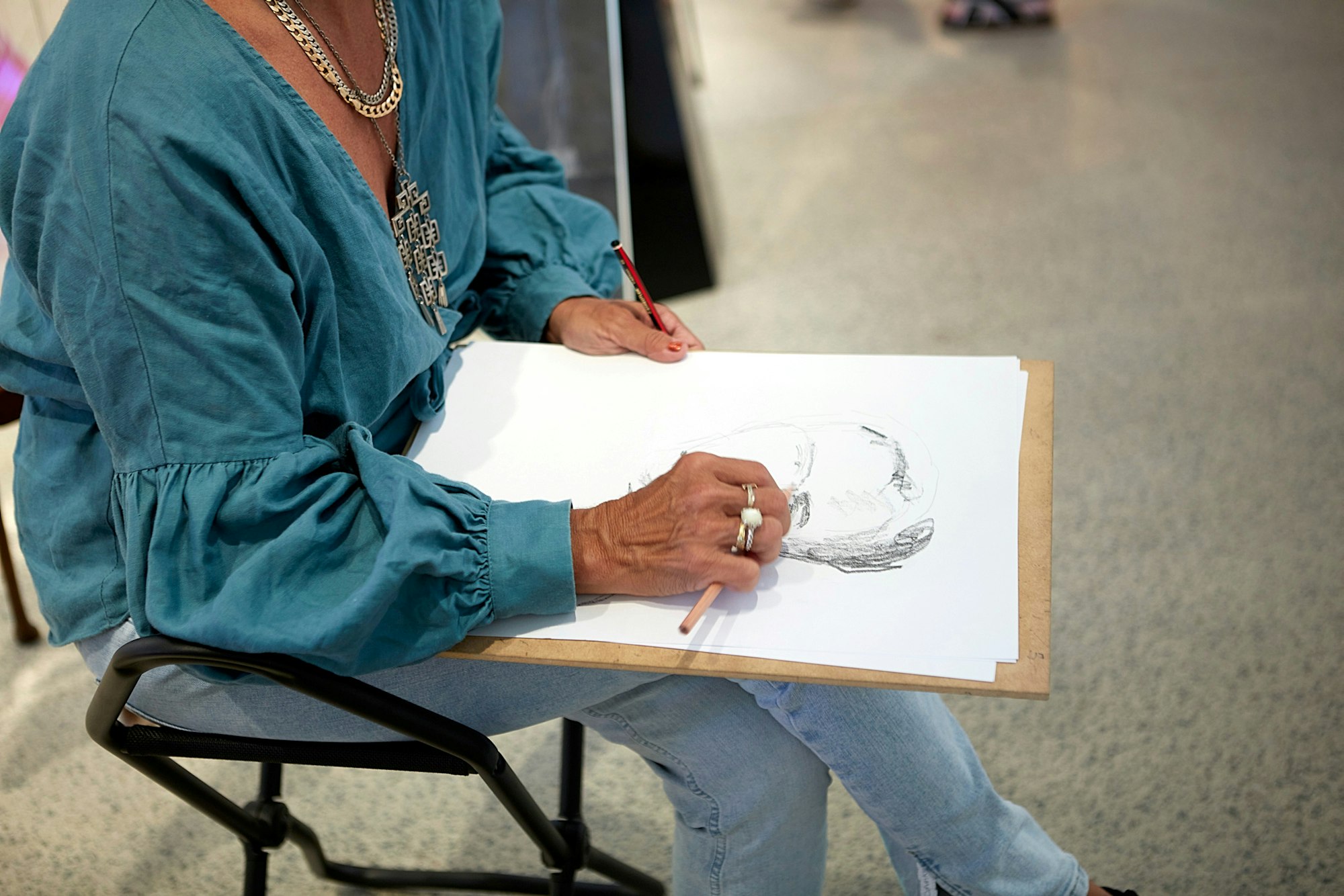 A seated person sketches with a pencil on paper, which rests on a board on their lap