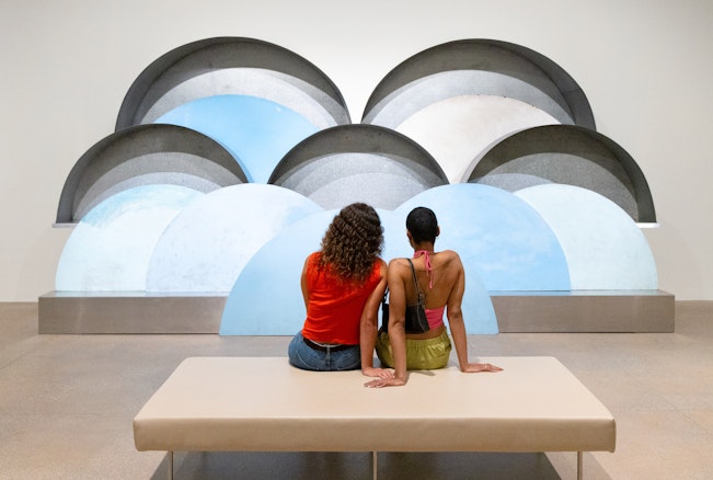 Two people sit looking at a large sculpture of overlapping cloud-like semi-circles