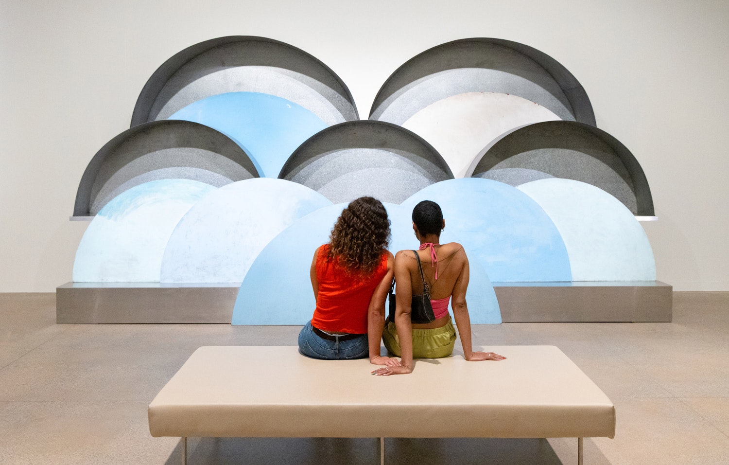 Two people sit looking at a large sculpture of overlapping cloud-like semi-circles