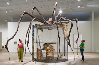 Two people in a gallery space looking at a large sculpture of a spider on top of a mesh enclosure