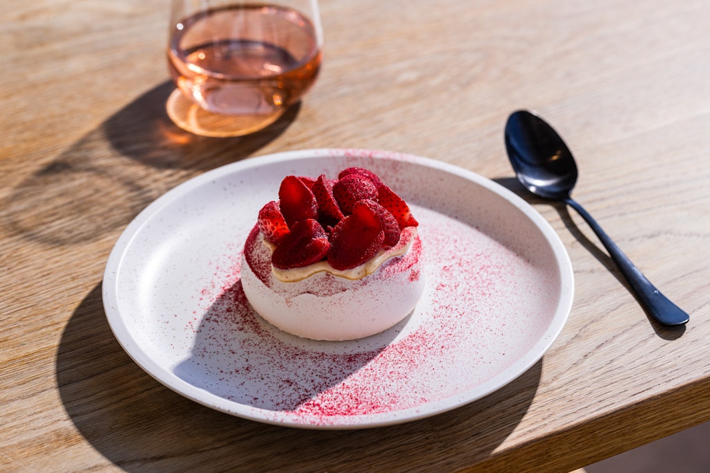 A strawberry-topped dessert on a place with a spoon and a glass of rosé alongside