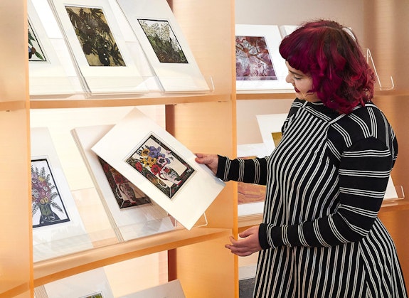 A person holds an art print in front of shelves of other prints