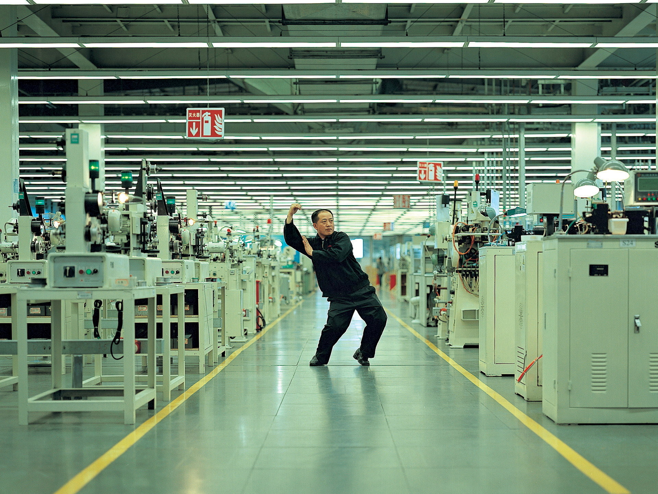 A person with short black hair and light skin dances in the middle of an enormous room full of industrial machines.