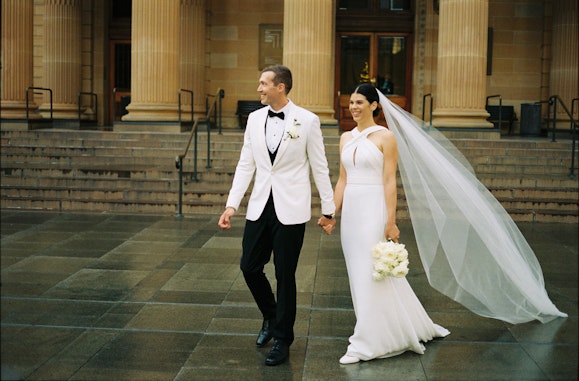 A person in a formal suit and another in a long white dress hold hands in front of a sandstone building