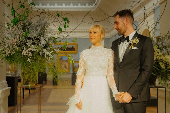 Two people stand holding hands in front of artworks and floral arrangements