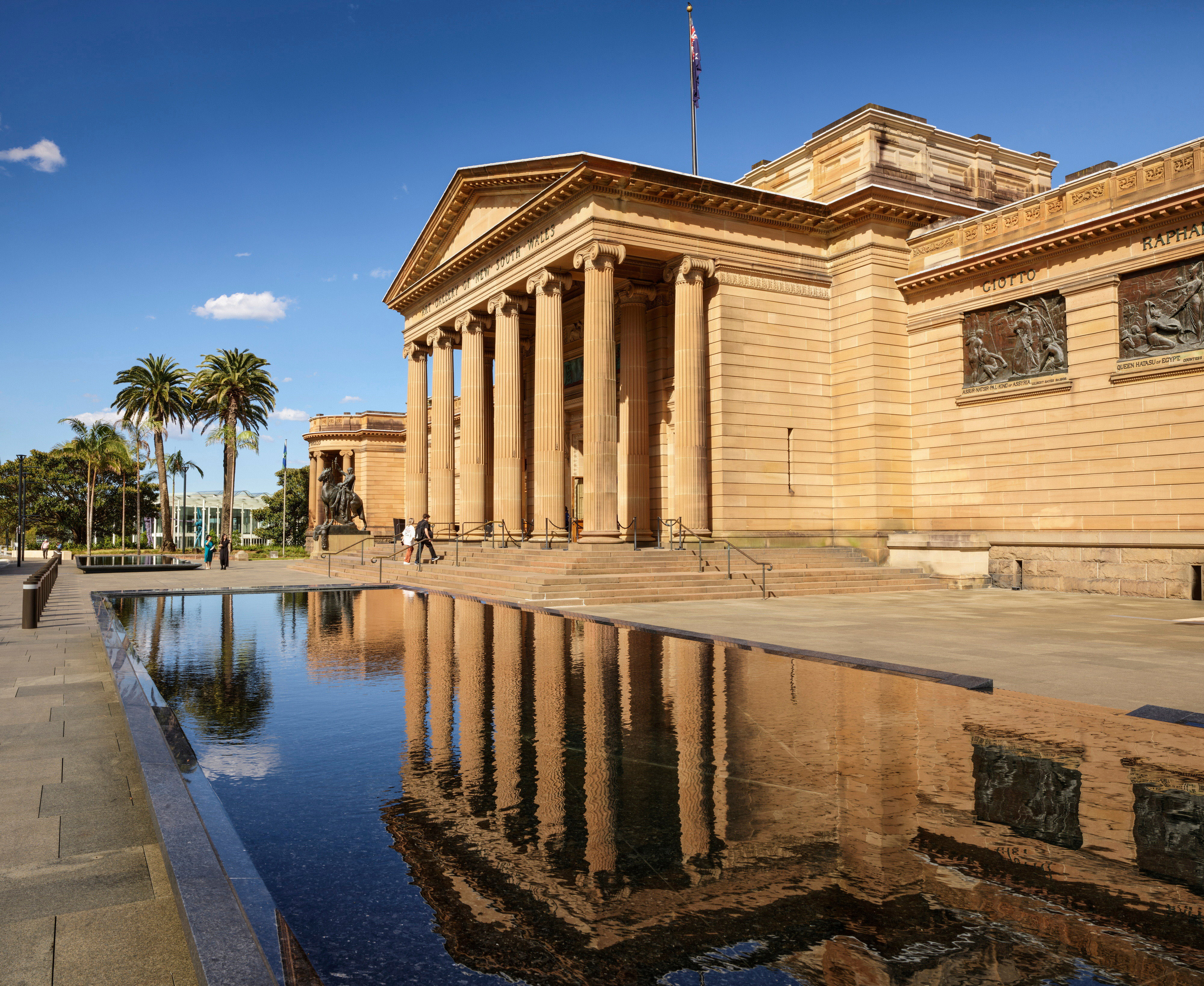 A sandstone building with a columned portico is reflected in a decorative pool