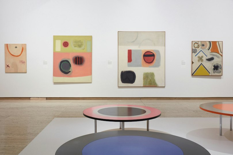 Brightly coloured abstract artworks hang on the gallery wall, while \ round sculptures are displayed in the centre of the room.