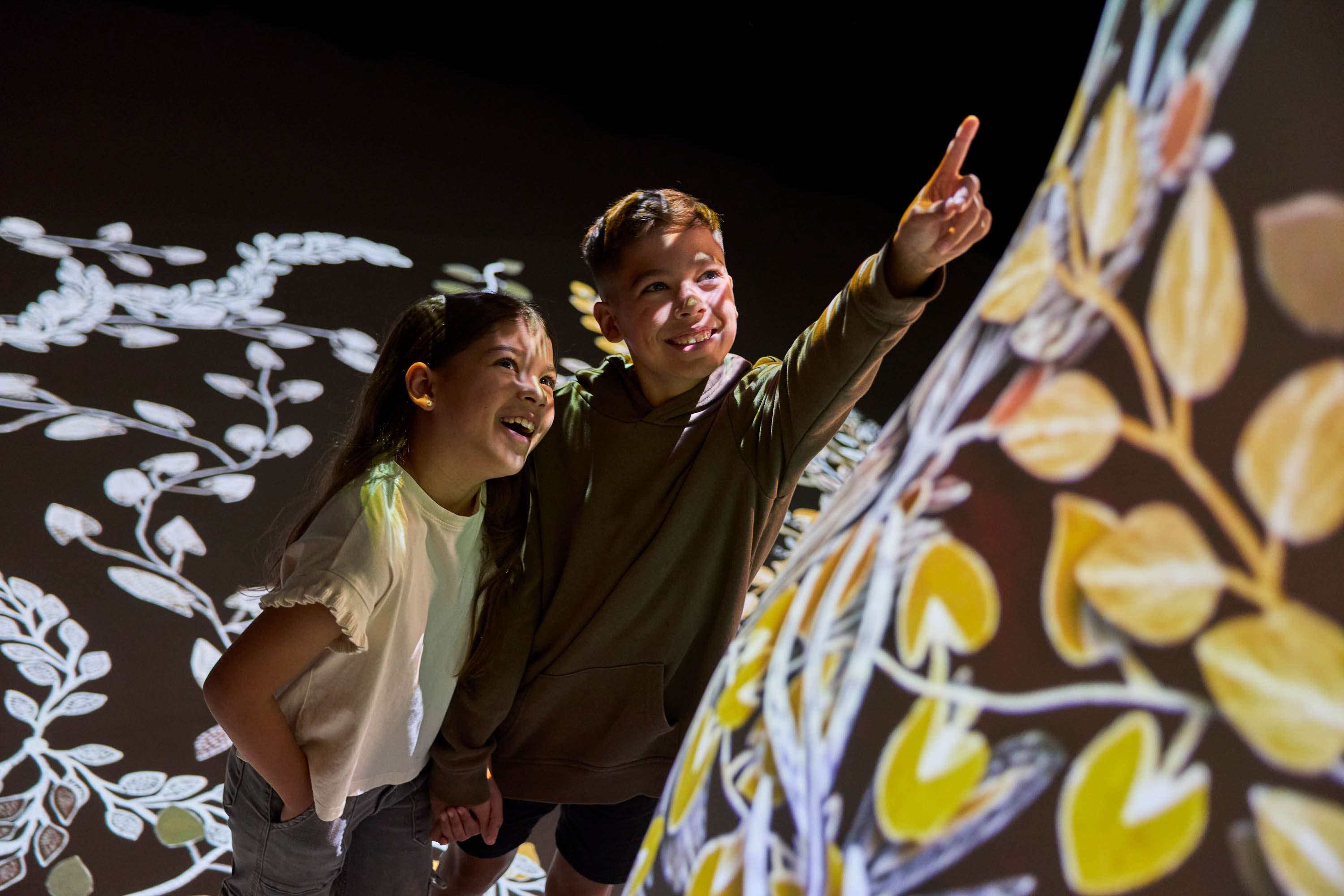 Two children smiling and pointing at an illuminated digital artwork with nature designs