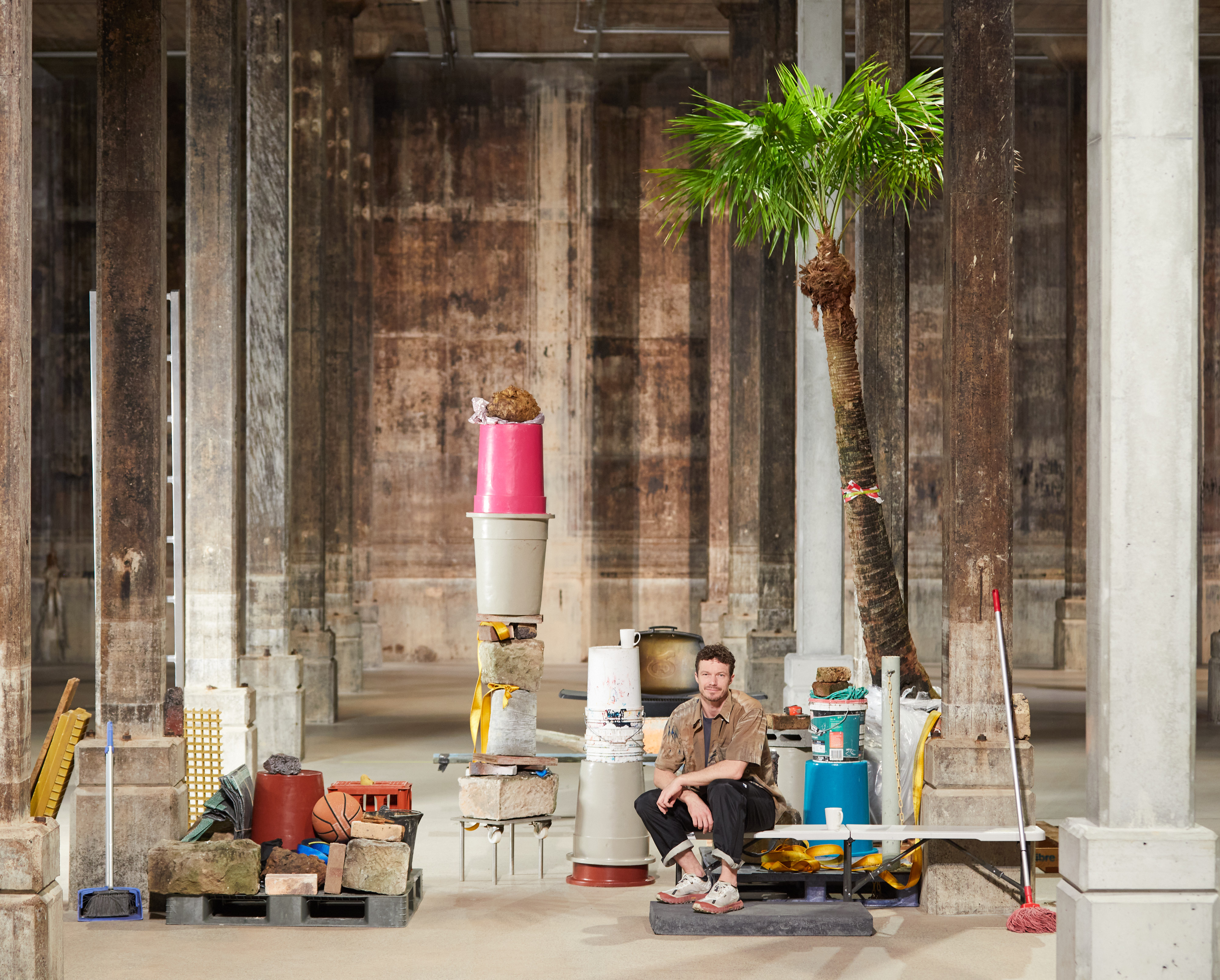 A man seated in a large cavernous space surrounded by assorted objects, including bricks, containers, a mop, and a palm tree.