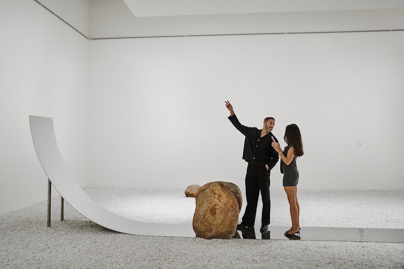 Two people standing on a curved mirrored surface among gravel and boulders
