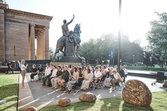 People gathered outside a sandstone building for a talk, with a statue of a man on a horse behind them