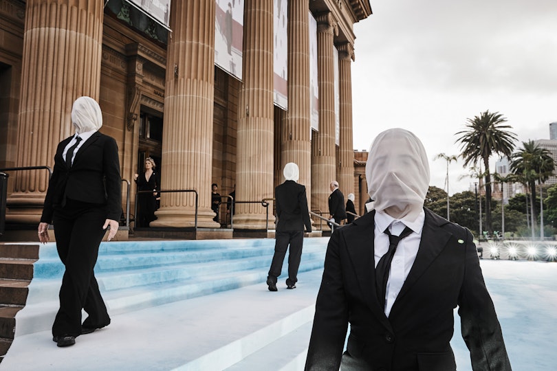 Performers in black suits with faces covered in white standing before a sandstone building