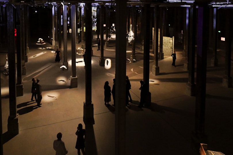 Visitors viewing artworks and sculptures in a large, dimly lit gallery with column