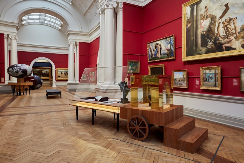 A red-walled gallery hung with paintings with a series of sculptural objects and display cases displayed on the wooden floor