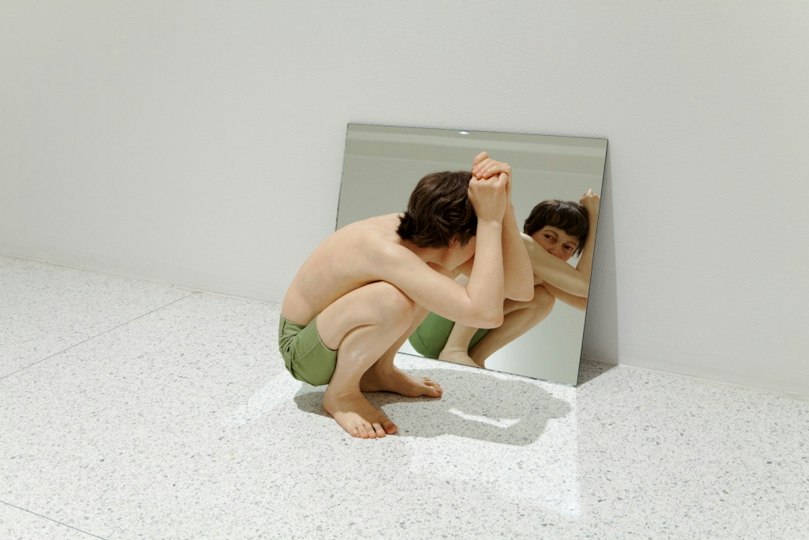 A lifelike sculpture of a boy in green shorts, squatting as he gazes into a mirror.