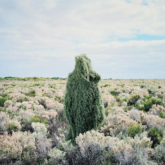 Person camouflaged in a green grass-like ghillie suit standing among dense shrubs in a landscape
