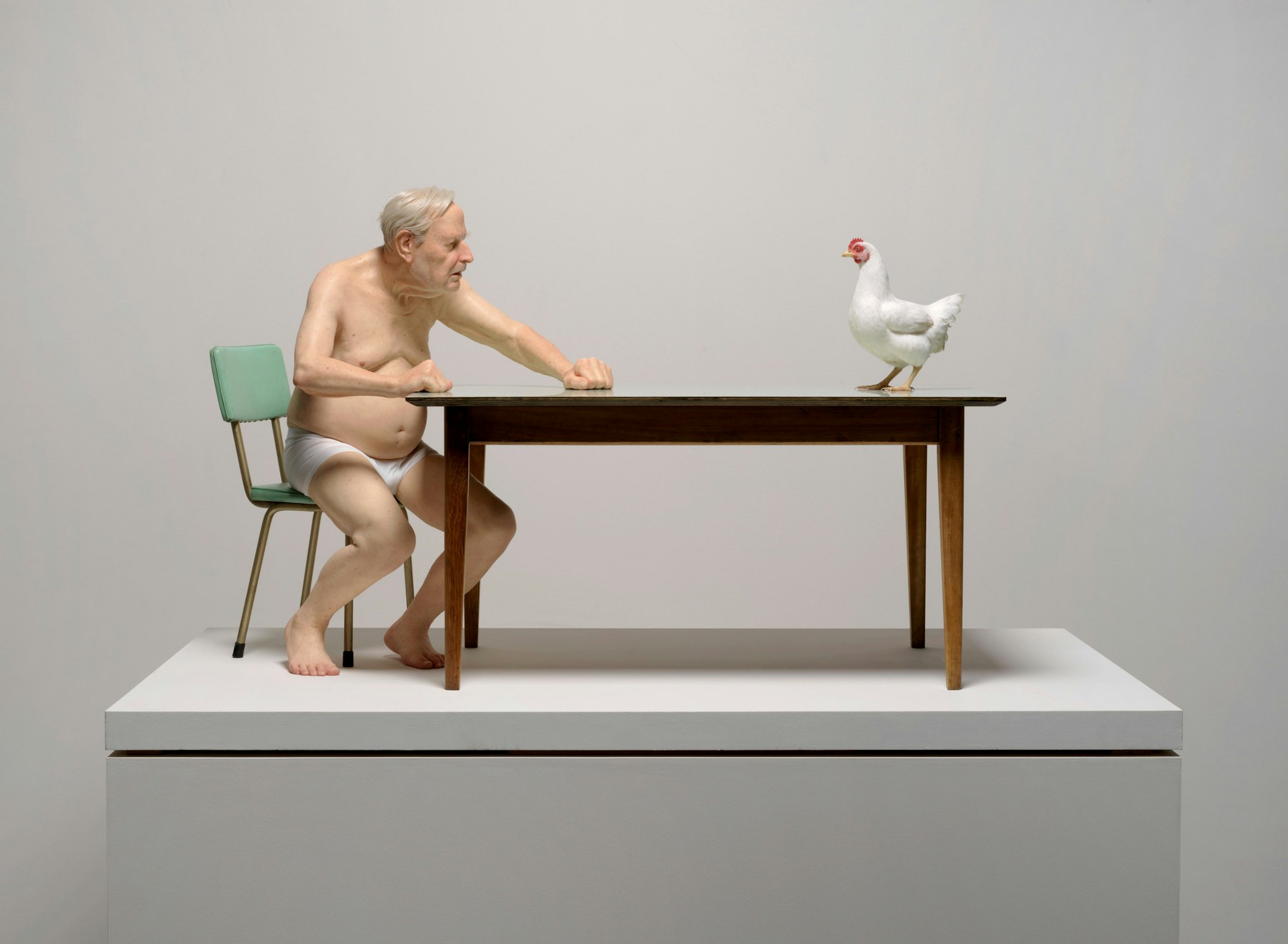 Lifelike sculpture of a man in underwear sitting at a table, facing a white chicken standing on the tabletop.