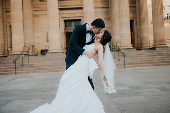 Two people embrace in front of a building. One person wears a suit. The other wears a white dress and carries a bouquet.