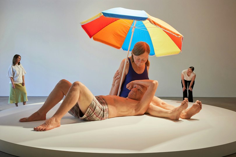 People looking at two larger-than-life sculptural figures under a beach umbrella