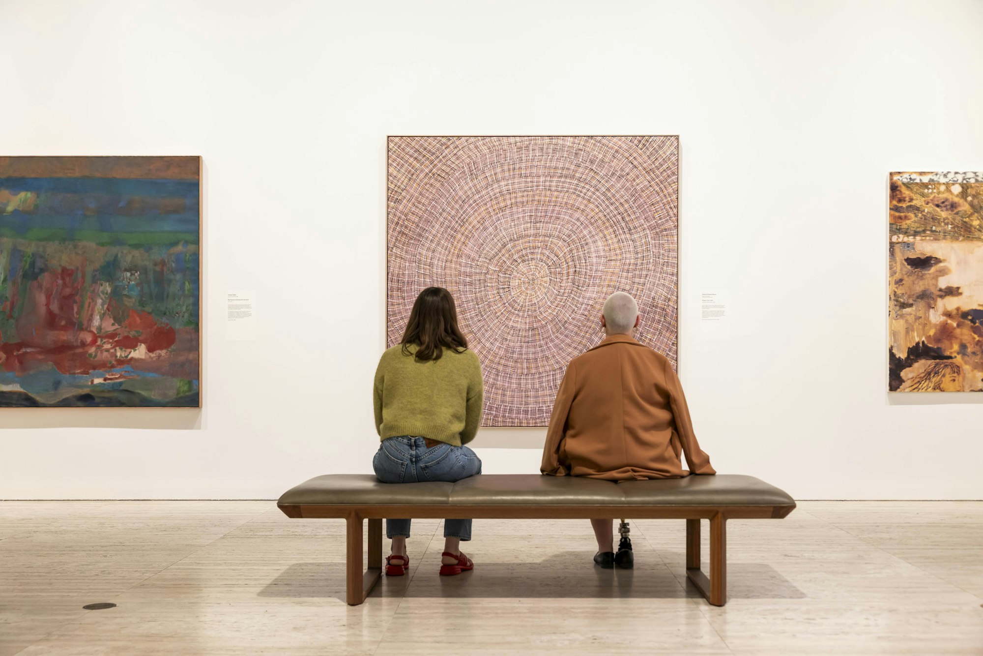Two people sit in an art gallery and view three large paintings on a wall