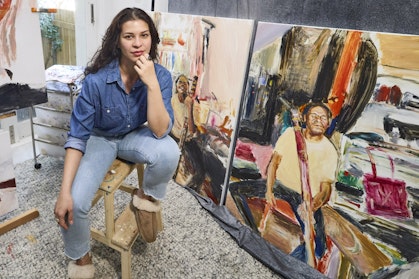 An artist in a light-filled studio surrounded by portrait paintings and paint tools