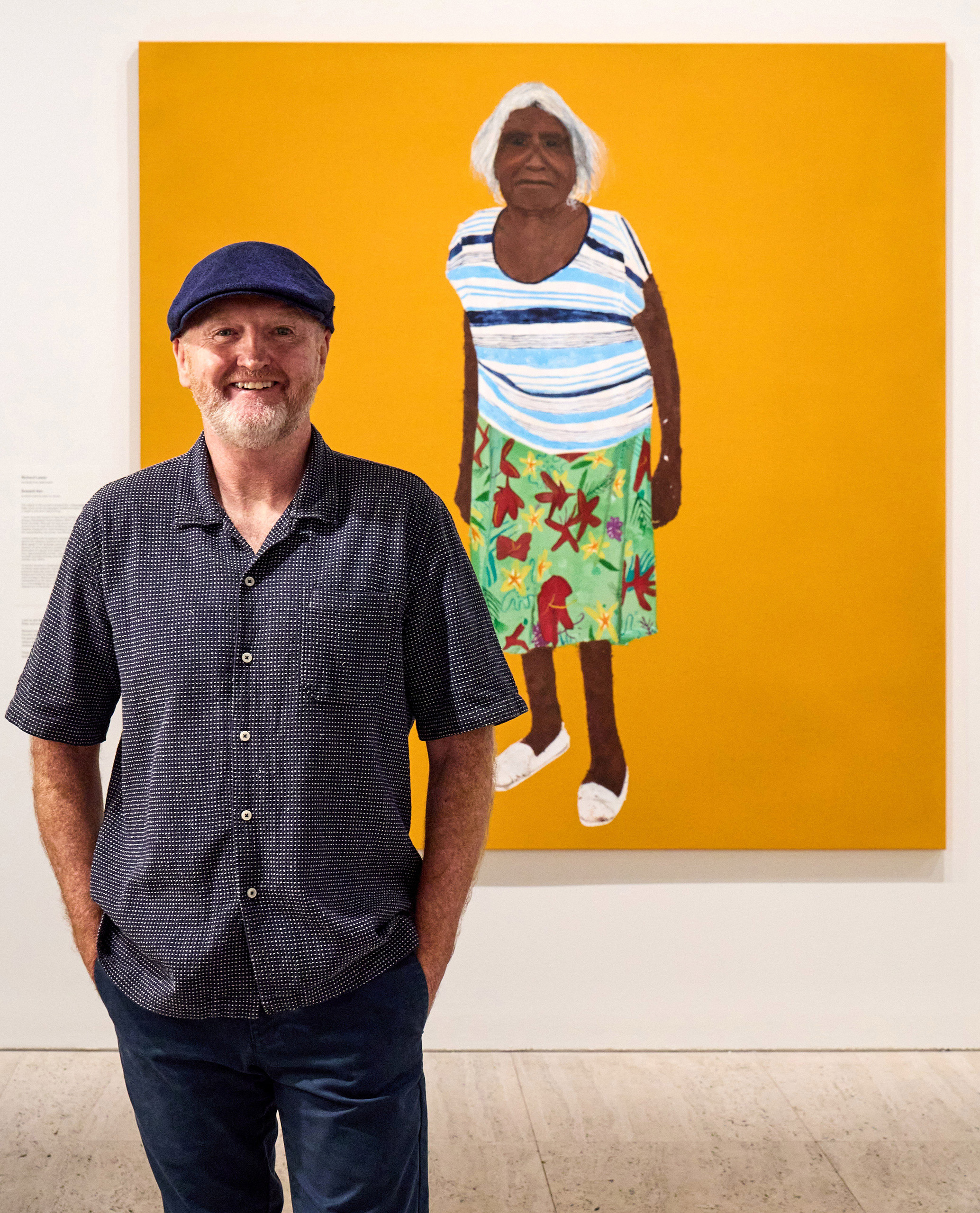 Smiling man in blue flat-cap hat stands in front of a portrait painting.