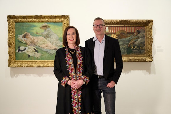 A man and woman standing in front of two paintings hung on a wall.