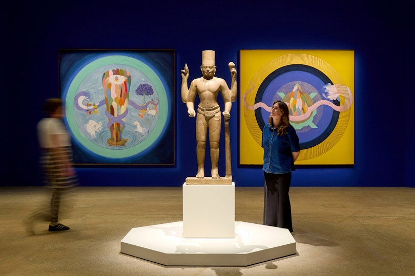 Two people view a statue and paintings on a blue wall in an art gallery