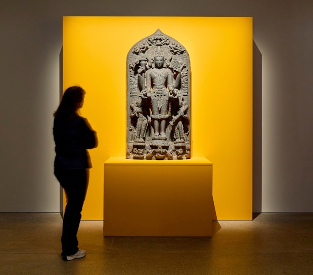 A person viewing an ancient sculpture displayed against a yellow wall.