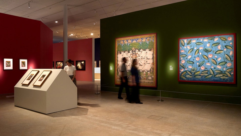People view paintings displayed on walls and plinths in a large gallery space.