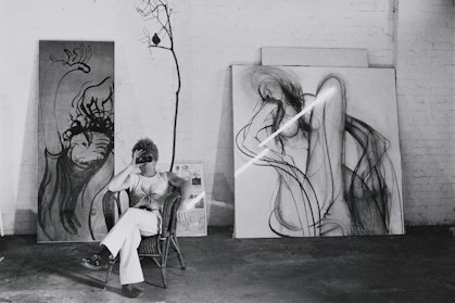 Black-and-white photo of a man in a cane chair looking through binoculars, with large paintings against an exposed brick wall behind him.