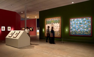 Visitors inside a gallery space looking at large paintings displayed on the walls.