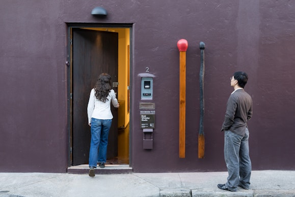 Two people at a building entrance beside a sculpture of two oversized wooden matchsticks, one burnt.