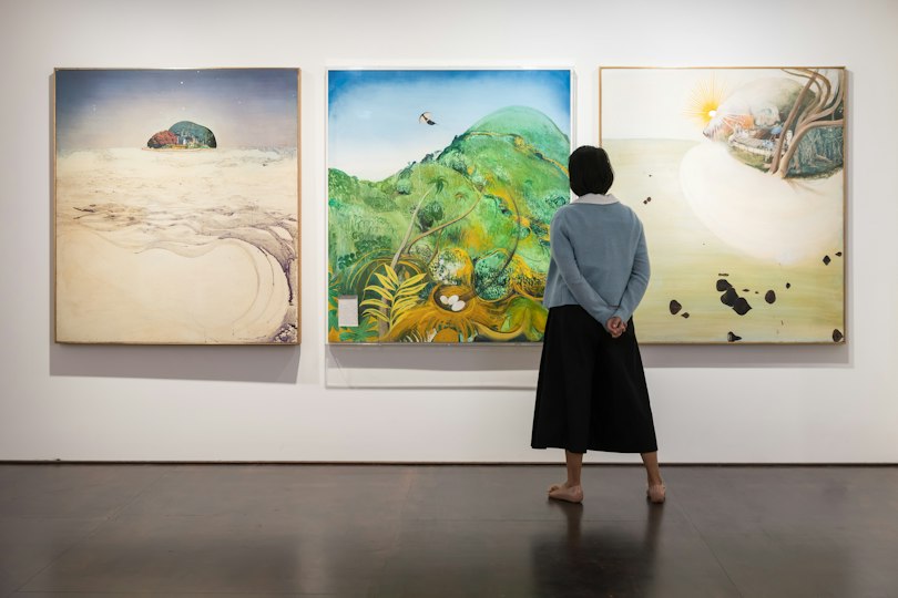 A woman stands in front of three landscape paintings displayed on a gallery wall.
