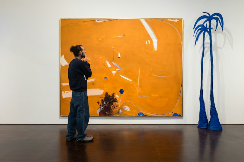 A man stands in front of an orange landscape painting and a blue palm tree sculpture displayed in an art gallery.