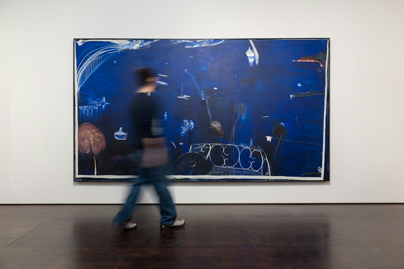 A blurred person walks past a large blue abstract painting displayed on a gallery wall.