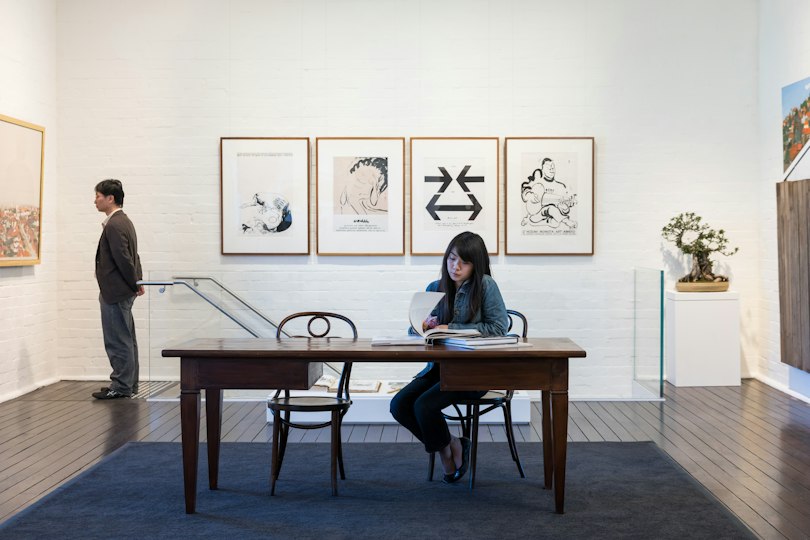 A man looks at a painting on a gallery wall while a woman seated at a table flips through a book, with artworks displayed in the background.