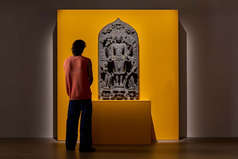 A visitor looks at an intricate Vishnu statue displayed against a vivid yellow gallery wall.