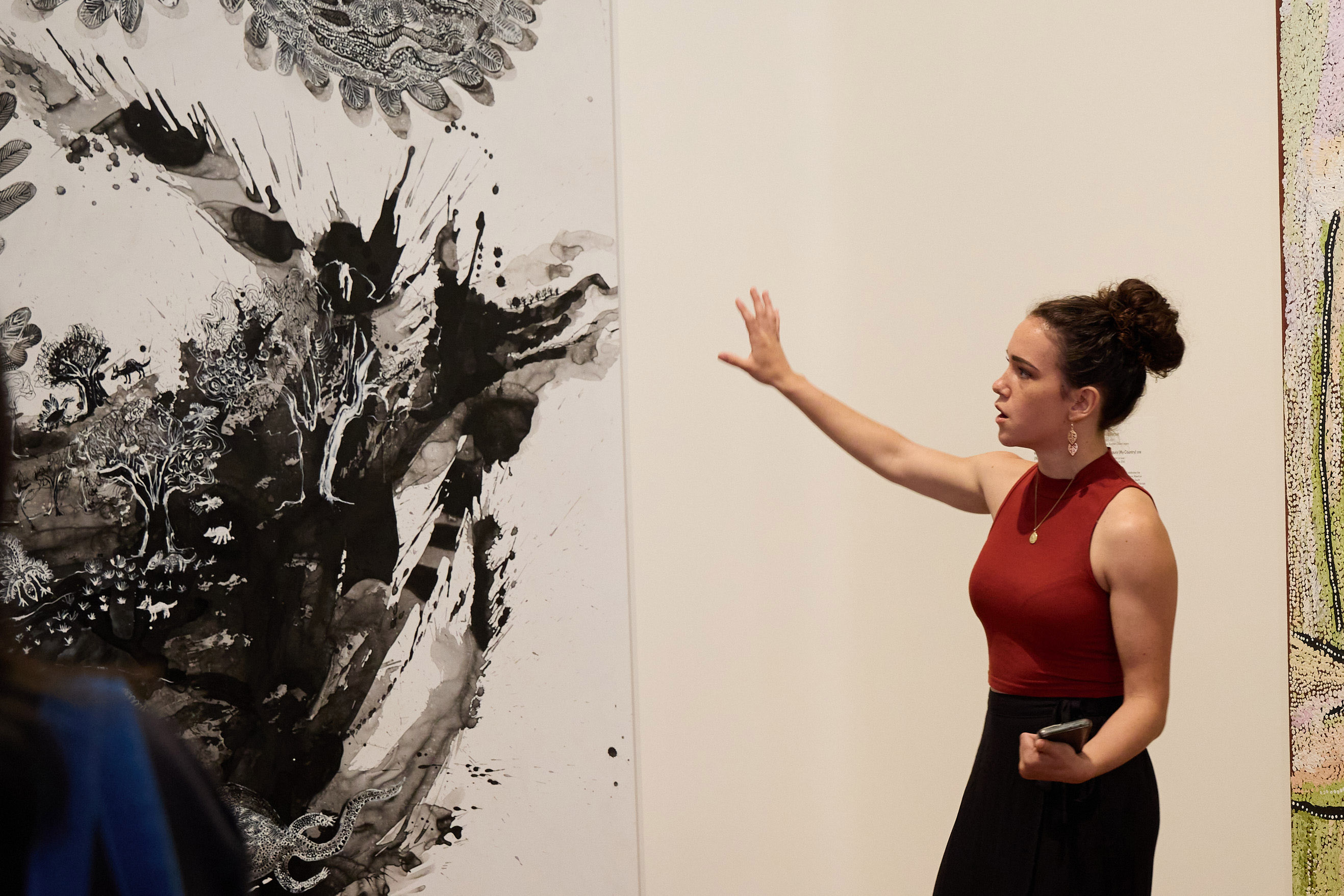 A woman gestures toward a large painting while leading a gallery tour.