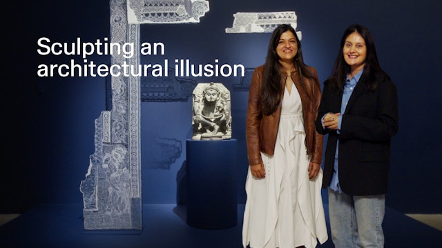 Two women standing in front of a sculpture and fabric artwork.