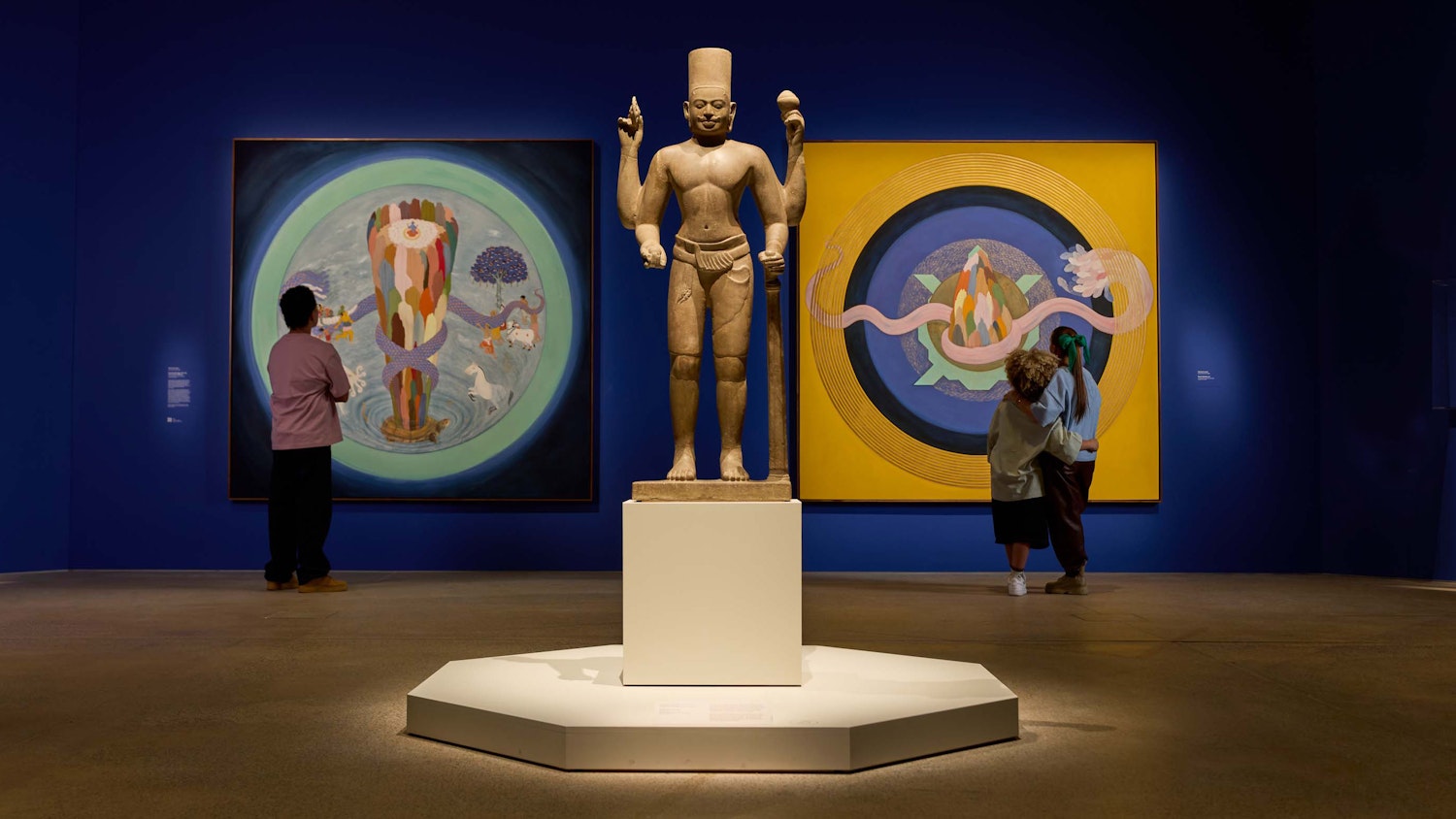 Two people view a statue and paintings on a blue wall in an art gallery