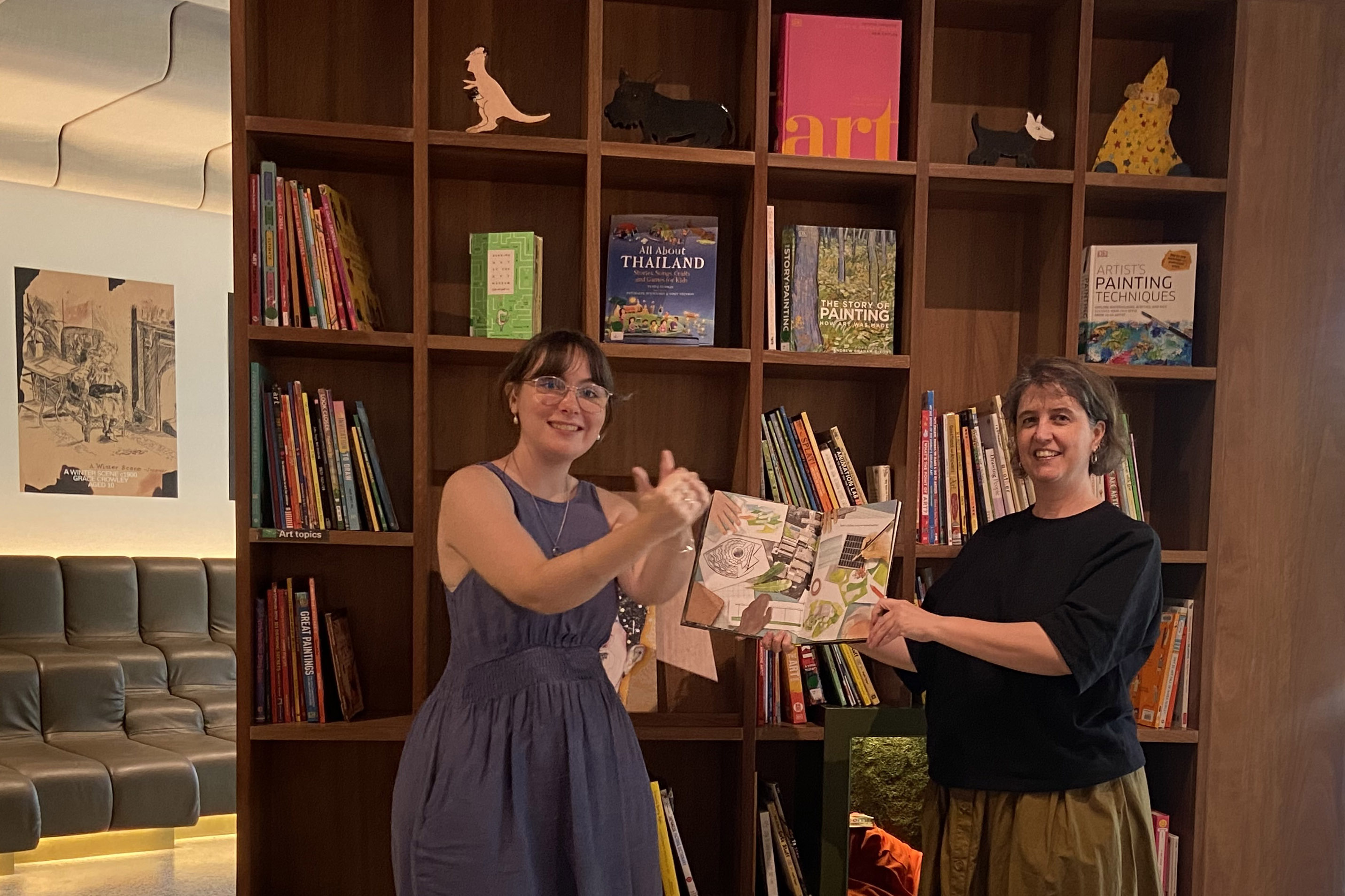 Two women stand in front of a book; one holds it while the other signs expressively.