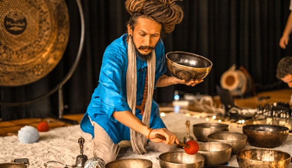A man seated on the floor, holds a sound bowl handle above various sound bowls in the foreground.