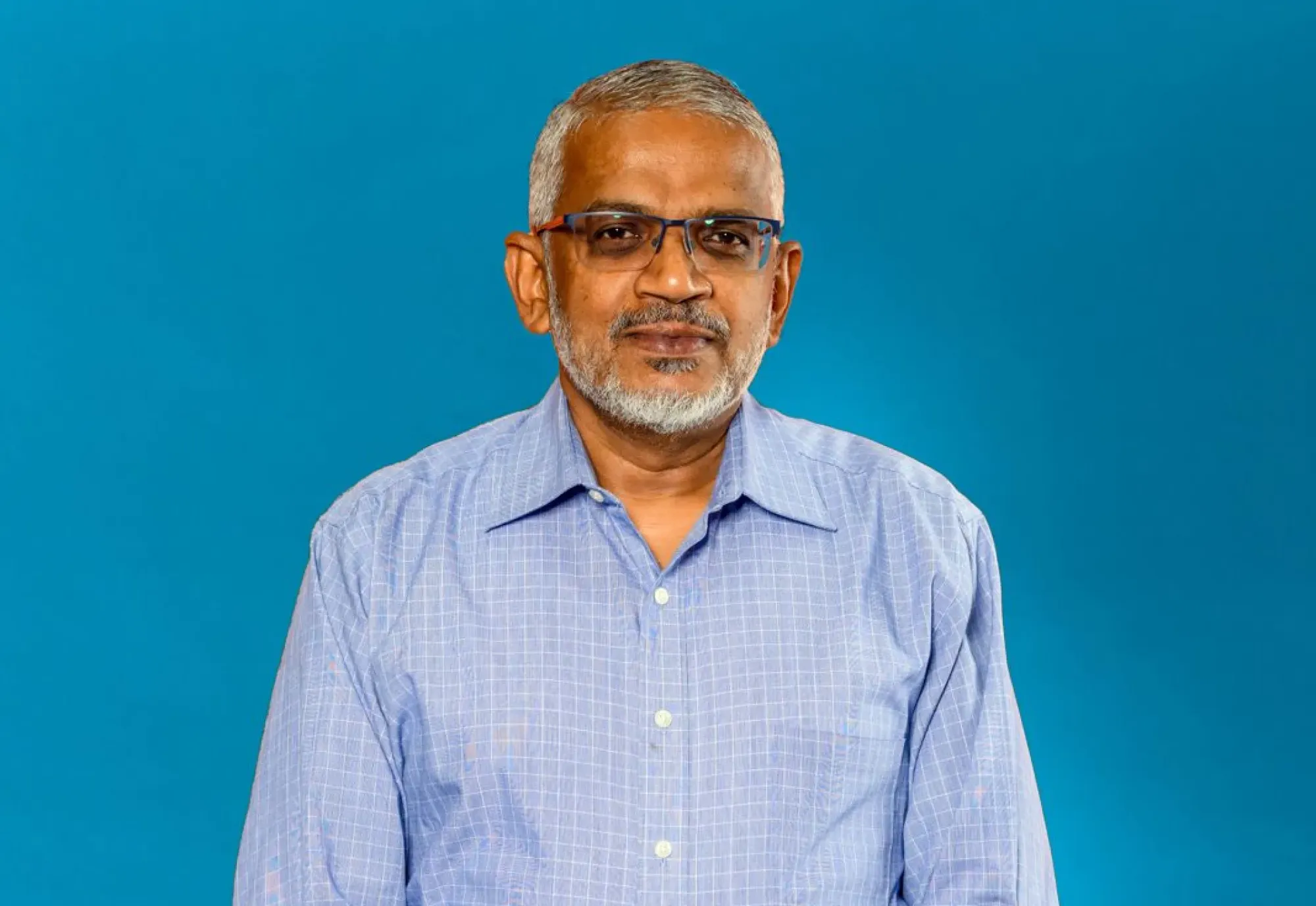 A man wearing glasses, standing in front of a blue background.
