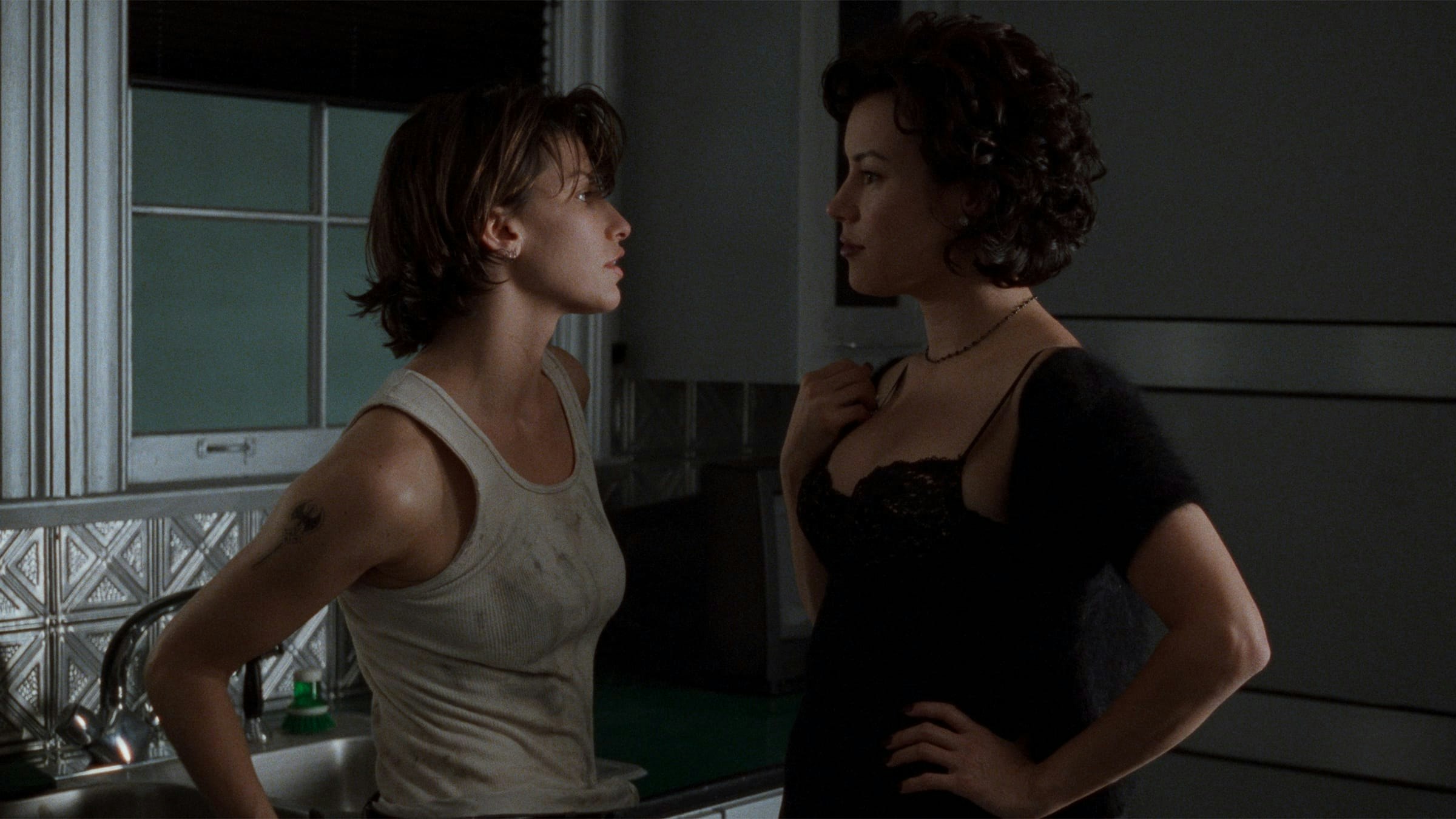 Two women staring at each other in a moodily lit kitchen.