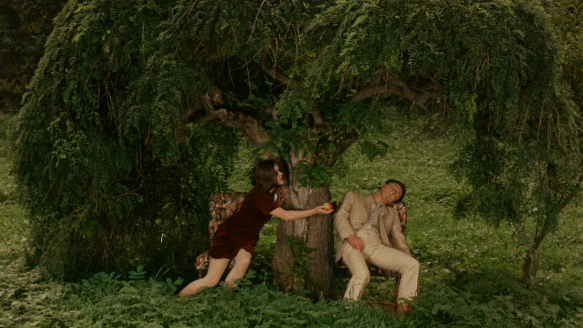 A woman offering an apple to a man in lush green woodland.